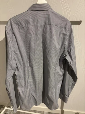 Randig skjorta från Riley, grå - Snygg grå skjorta från Riley med tunna vita ränder och klassisk krage. Skjortan har svarta knappar och lång ärm, samt ett diskret mönstrat tyg på insidan av knappslån. Perfekt för dig som gillar stilrena detaljer och en clean look.