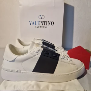 Valentino Garavani vita sneakers 43 - Snygga vita sneakers från Valentino Garavani med bred svart panel på sidan och coola nitar vid hälen. Skorna har snörning och en platt sula med rutmönster undertill. Tillverkade i skinn för en lyxig känsla och stilren look. Låda ingår ej.