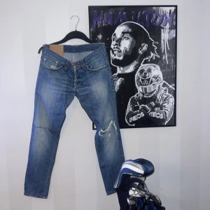 Blå jeans från dondup - Säljer ett par blå jeansbyxor från dondup med coola slitningar och detaljer på både fram- och baksida. Klassisk femficksmodell med kontrastsömmar och tryckt logga på linningen bak. Perfekta för dig som gillar en avslappnad och trendig look.