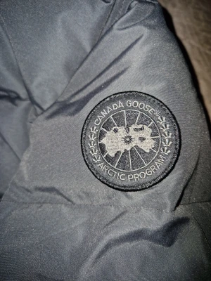 Mörkgrå pufferjacka från Canada Goose - Säljer en mörkgrå pufferjacka från Canada Goose, modell Fusion M/M. Jackan har huva, ribbade muddar och snyggt broderat logomärke på ärmen. Fylld med dun och har dragkedja framtill. Perfekt för kalla dagar och riktigt stilren look.