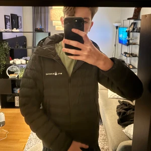 Mörkgrön dunjacka från Sail Racing - Mörkgrön  dunjacka från Sail Racing med huva och diskret logga på bröstet. Jackan är quiltad, har dragkedja framtill och är fylld med 800 fill power dun. Perfekt för kalla dagar och har en clean, sportig look.