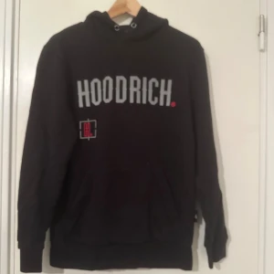 Svart hoodie från Hoodrich med tryck - Svart hoodie från Hoodrich i storlek small med stor logotyp i grått och rött framtill och på ryggen. Tröjan har huva med snörning, känguruficka och coolt street-inspirerat tryck på både fram- och baksida samt text på ärmen. Perfekt för dig som gillar urban stil.