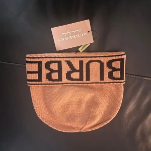 Beige Burberry stickad mössa - Snygg beige stickad mössa från Burberry med bred uppvikt kant och svart logotyp i grafisk stil runt hela kanten. Perfekt för dig som vill ha en trendig accessoar med lyxig känsla och tydlig branding.