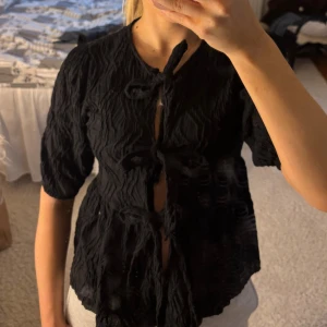 Svart blus med knyt från Gina Tricot - Säljer en svart blus från Gina Tricot i storlek XS. Blusen har korta ärmar, crinklat tyg och flera snygga knyt framtill. Säljer då den ej kommer till användning 💞