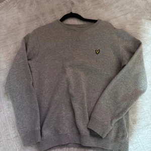 Grå sweatshirt från Lyle & Scott - Säljer en stilren grå sweatshirt från Lyle & Scott med deras klassiska gula fågel-logga på bröstet. Tröjan har rund hals, ribbade muddar och är långärmad. Perfekt för dig som gillar enkel och snygg streetstil.