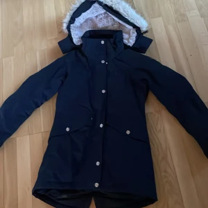 Mörkblå vinterjacka från 8848 Altitude - Snygg mörkblå parkajacka från 8848 Altitude med silverfärgade knappar och huva med vit fuskpäls. Jackan har långa ärmar, två stora fickor framtill och broderad logga på bröstet och ärmen. Perfekt för kalla dagar och har en stilren look.