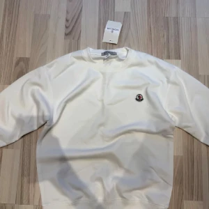 Moncler Tröja - Vit sweatshirt från Moncler med rund hals och diskret logotyp-patch på bröstet. Tröjan har ribbade muddar vid hals, ärmslut och nederkant. Tillverkad i mjuk bomull som känns skön mot huden. Perfekt för dig som gillar clean och stilren look.