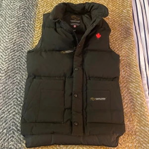 Svart dunväst från Canada Goose - Svart dunväst från Canada Goose med hög krage, två stora fickor framtill och broderad röd lönnlöv på bröstet. Västen har både dragkedja och tryckknappar samt märkets logga på fickan och insidan. Perfekt för kalla dagar och snygg att styla över hoodie.