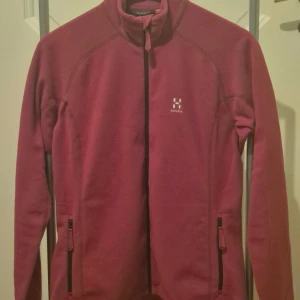 Fuchsia fleecejacka från Haglöfs L - Säljer en fuchsia fleecejacka från Haglöfs i storlek L. Jackan har hel dragkedja framtill, två fickor med dragkedja och en hög krage med snörning. Perfekt för kyliga dagar och outdoor-aktiviteter. Stilren design med diskret Haglöfs-logga på bröstet.