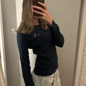 Mörkblå kabelstickad tröja bondelid  - Säljer en mörkblå kabelstickad tröja från bondelid med broderad logga på bröstet. Tröjan har rund halsringning, ribbade muddar och lång ärm. Perfekt för dig som gillar klassisk och stilren look.