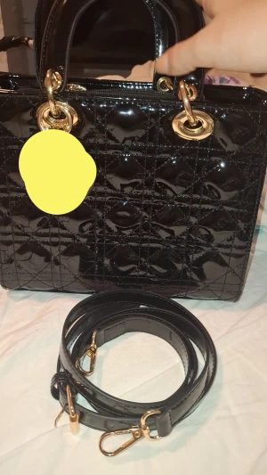 Svart quiltad handväska  - Snygg svart handväska i glansigt skinn med quiltat mönster och guldiga metalldetaljer. Väskan har både handtag och avtagbar axelrem, perfekt för dig som vill ha en lyxig och stilren accessoar. Klassisk boxig form och dragkedja upptill.