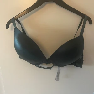 Svart bh från Censored 90D - Svart bh från Censored i storlek 90D med vadderade kupor och justerbara axelband. Sidorna har spetsdetaljer som ger en snygg touch. Perfekt för dig som vill ha både stöd och stil.