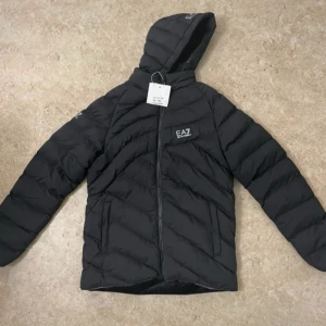 Svart pufferjacka från EA7 - Svart pufferjacka från EA7 med huva och snygga diagonala quiltade sömmar. Jackan har dragkedja framtill och två sidofickor. EA7-logga på bröstet och ärmen ger en sportig vibe. Perfekt för kalla dagar när du vill hålla stilen.