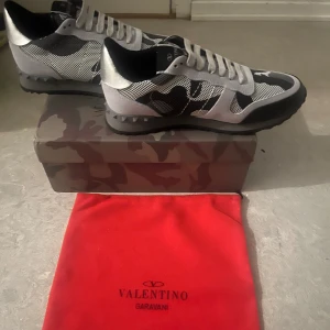 Valentino Rockrunner sneakers svart/grå - Snygga Valentino Rockrunner sneakers i svart och grått med coolt kamouflagemönster. Skorna har snörning, platt sula och detaljer i mocka och mesh. Ikoniska nitar på hälen och en rund tå ger en modern vibe. Perfekt för dig som gillar exklusiva streetwear.