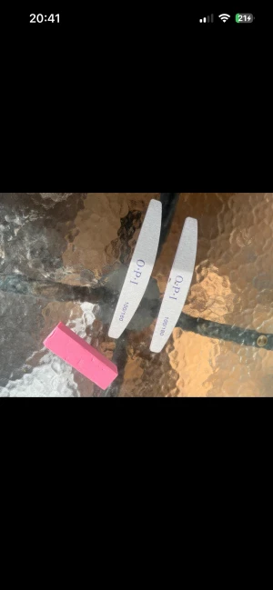 Nagelfilar och buffert från OPI - Två vita nagelfilar från OPI med lila text och en rosa fyrkantig nagelbuffert. Perfekt för att fixa snygga naglar hemma och forma dem precis som du vill. Filarnas form är böjd och bufferten är rektangulär, båda i slitstarkt material. Nya endast provad 