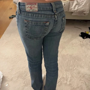True Religion blå bootcut jeans - Säljer ett par blå jeans från True Religion med klassiska kontrastsömmar och snygga detaljer på bakfickorna. Modellen är lågmidjad med bootcut och har slitningar nertill för en cool vibe. Jeansen är i mjukt denimtyg och har logga på bakfickan.
