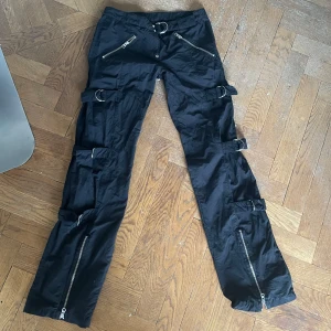 coola svarta byxor med spännen - Perfecta byxor för t.ex ett rave!! De är lågmidjade, bootcut eller baggy/raka som du själv kan bestämma genom att ändra på spännena och dragkedjan längst ner. Byxorna är i typ samma material som parachute pants. Tyvärr var dessa as snygga byxor för stora för mig så jag hoppas att dem kommer till någon som kan njuta av dom lika mycket som jag önskar att jag kunde🫶