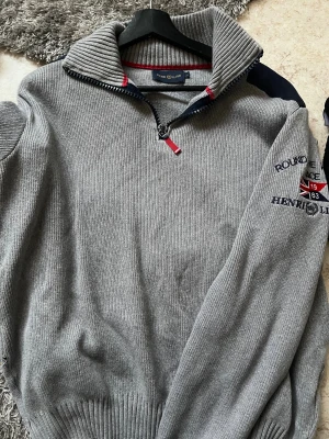 Grå stickad tröja från Henri Lloyd - Snygg grå stickad tröja från Henri Lloyd med broderad logga och flagga på ärmen. Tröjan har en ribbad krage med half zip och röda detaljer vid dragkedjan. Perfekt för dig som gillar klassisk stil med en sportig touch.