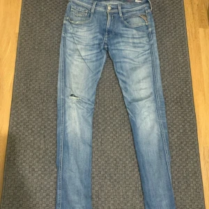 Replay Anbass ljusblå jeans slim fit - Replay Anbass jeans i ljusblå tvätt med snygga slitningar och en liten reva på ena benet. Slim fit-modell med klassiska fem fickor, dragkedja och Replay-logga på fickan. Perfekt för dig som gillar en modern och avslappnad stil.