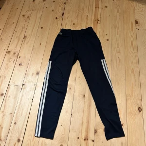 Mörkblå träningsbyxor Lacoste Sport - Snygga mörkblå träningsbyxor från Lacoste Sport med klassiska vita ränder längs benen och elastisk midja. Byxorna har en smal passform och är gjorda i mjukt syntetmaterial, perfekt för sport och chill. Crocodile-logga på höften för extra stilpoäng.