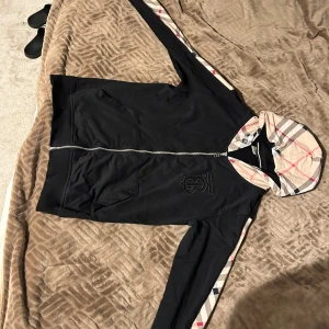 Svart Burberry kofta med rutig huva - Svart kofta från Burberry med klassisk rutig huva och detaljer längs ärmarna. Koftan har dragkedja framtill, fickor och ett broderat Burberry-emblem på bröstet. Rutigt mönster i beige, svart och rött ger en snygg kontrast till den svarta basen.