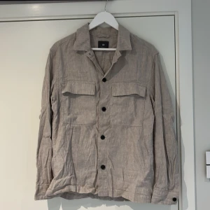 Beige overshirt från H&M i linne - Snygg beige overshirt från H&M i regular fit, tillverkad i linne. Skjortan har klassisk krage, svarta knappar framtill och två stora bröstfickor. Långärmad modell med knapp vid ärmslut.