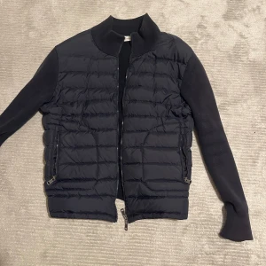 Moncler cardigan  - Snygg svart dunjacka från Moncler med quiltad framsida och stickade ärmar. Jackan har ribbad krage, två dragkedjeförsedda fickor och Moncler-logga på ärmen. Materialet är en mix av dun och stickad polyester, perfekt för kyliga dagar kan gå ner precis i snabb affär
