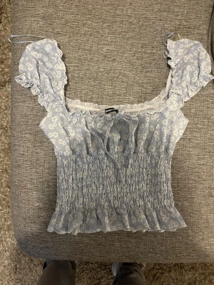 Blommig blå blus från Gina Tricot - Supersöt ljusblå blus med blommigt mönster från Gina Tricot. Toppen har volangdetaljer vid axlar och nederkant, smockad midja och en liten knytning framtill. Ärmlös design med breda axelband och romantisk vibe, perfekt för vår och sommar.