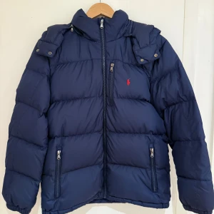 Mörkblå pufferjacka Polo Ralph Lauren  XL  - Snygg mörkblå pufferjacka från Polo Ralph Lauren i storlek XL (163-174 cm). Jackan har huva, flera dragkedjefickor och det klassiska röda Polo-logot broderat på bröstet. Tillverkad i polyester med quiltad design och elastiska muddar vid ärmslut. Perfekt för kalla dagar. Hängaren är trasig och lite skavt på muddarna (se bild). Jackan är nytvättad på kemtvätt så helt ren! 