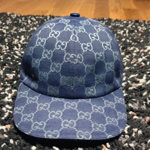 Blå Gucci keps med GG-mönster - Snygg blå keps från Gucci med det klassiska GG-mönstret över hela kepsen. Kepsen har böjd skärm och är tillverkad i bomullsmaterial med beige insida. Perfekt för dig som vill ha en exklusiv och trendig look.