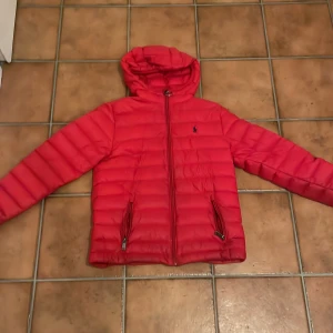Röd pufferjacka från Polo Ralph Lauren - Snygg röd pufferjacka från Polo Ralph Lauren med huva och klassisk logga broderad på bröstet. Jackan har två fickor med dragkedja. Perfekt för kalla dagar och har en modern, quiltad look. Defekter finns på bild 4 och 5 därav priset!