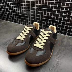 Maison margiela sneakers  - Snygga sneakers i mörkbrunt med svarta paneler och beige detaljer. Ovandelen är i mocka och skinn, med kontrasterande ljusa snören och gummisula i naturfärg. Rund tå och klassisk retrostil som passar perfekt till jeans eller cargo pants.
