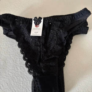 Svarta stringtrosor 4stycken. - Snygga svarta stringtrosor 4 stycken med spetsdetaljer och dekorativ rosett framtill.Byxorna har en feminin design med vågiga kanter och genombrutet mönster i spetsen. Perfekt för dig som gillar detaljer och stil.