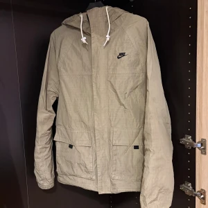 Beige vindjacka från Nike med huva - Säljer en beige vindjacka från Nike med huva och snörning. Jackan har två stora fickor framtill, svart Nike-logga på bröstet och dragkedja. Materialet är lätt och syntetiskt, perfekt för blåsiga dagar. Passformen är normal och jackan är långärmad.