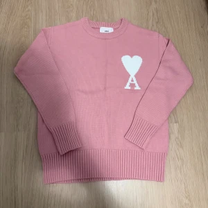 Rosa stickad tröja från Ami - Säljer en rosa stickad tröja från Ami Alexandre Mattiussi med vit hjärt- och A-logga på bröstet. Tröjan har rund halsringning, ribbade muddar och är långärmad. Perfekt för dig som vill ha en snygg och trendig look. Size S