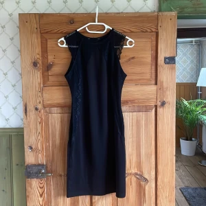 Svart spetsklänning med ärmlös design - Snygg svart klänning med spetsdetaljer längs sidorna och axlarna. Klänningen är ärmlös och har en rak, smickrande passform som ger en elegant siluett. Perfekt för dig som gillar stilrena plagg med en twist av spets.