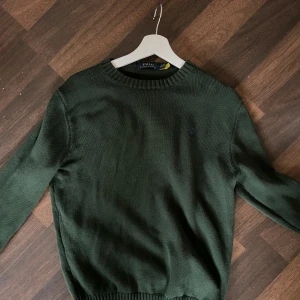 Grön stickad tröja från Polo Ralph Lauren - Mörkgrön stickad tröja från Polo Ralph Lauren. Tröjan har rund hals, ribbade muddar och den klassiska broderade loggan på bröstet. Perfekt för dig som gillar stilrena och tidlösa plagg med en chill vibe. Tröjan är storlek M men skulle nog säga att den passar S mer!