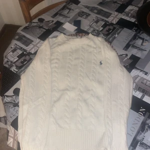 Vit kabelstickad tröja från Polo Ralph Lauren - Säljer en vit kabelstickad tröja från Polo Ralph Lauren med klassisk rund hals och blå broderad logga på bröstet. Tröjan har långärmade ribbade muddar och är gjord i mjuk bomull, perfekt för dig som gillar clean och stilren look.