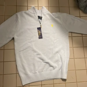 Ljusblå stickad half zip från Polo Ralph Lauren - Snygg ljusblå stickad tröja från Polo Ralph Lauren med half zip och ribbad krage. Klassisk gul logga broderad på bröstet. Tröjan har långärmad passform och är perfekt för dig som gillar stilrena och fräscha plagg.