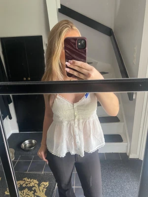 Vit blus med spets och knappar - Säljer en söt vit blus med tunna axelband, spetsdetaljer och knappar framtill. Blusen har en lätt v-ringning och volangkant nertill. Perfekt för varma dagar och ger en romantisk vibe.