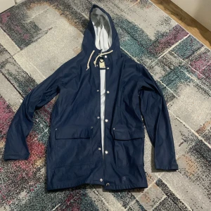 Mörkblå regnjacka med huva - Snygg mörkblå regnjacka från Tretorn med vit insida och huva med snörning. Jackan har två stora fickor framtill, tryckknappar och en reflexdetalj baktill. Perfekt för blöta dagar och har en clean, stilren look.