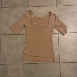 Beige topp med djup urringning - Säljer en beige topp med djup rund urringning och halvlånga ärmar. Modellen är figurnära och har en enkel, stilren design utan mönster eller detaljer. Perfekt att styla med jeans eller kjol för en clean look.