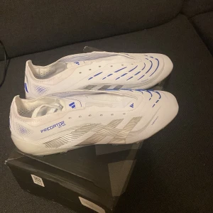 Adidas Predator fotbollsskor vit/blå - Säljer ett par Adidas Predator elite fotbollsskor i vitt med blå detaljer och silvermönster. Skorna har låg profil, platt sula med dobbar och en snygg, modern design. Tillverkade i syntetmaterial med ventilerande meshpartier och Predator-logga på sidan och sulan.