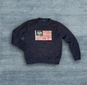 Mörkblå stickad tröja Polo Ralph Lauren - Mörkblå stickad tröja från Polo Ralph Lauren med rund halsringning och lång ärm. På bröstet finns en stor broderad patch i amerikansk flaggstil med text och RL-detaljer. stil