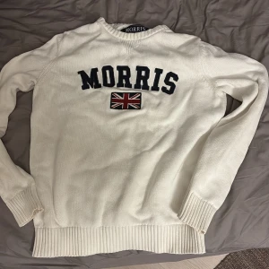Vit stickad tröja från Morris - Vit stickad tröja från Morris med broderad logga och brittisk flagga framtill. Tröjan har rund hals, långa ärmar och breda ribbade muddar vid ärmslut och nederkant. Perfekt för en klassisk och stilren look.