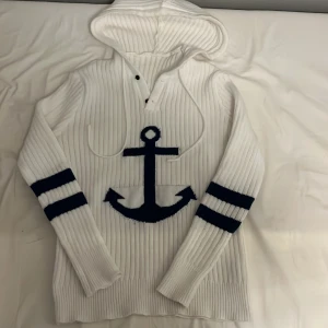 Vit stickad hoodie  - Vit ribbstickad hoodie med huva och snörning, prydd med ett stort marinblått ankare på bröstet och två marinblå ränder på varje ärm. Långärmad och perfekt för en chill och nautisk stil.