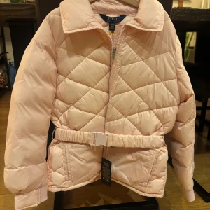 Ljusrosa quiltad pufferjacka Polo - Snygg ljusrosa pufferjacka från Polo Ralph Lauren med quiltat mönster och dragkedja framtill. Jackan har ett justerbart bälte i midjan, två fickor med dragkedja och ribbade muddar vid ärmslut. Perfekt för kyliga dagar och ger en cool vibe. Nypriset  är 1995kr och nu säljer jag den för 900kr, priset kan diskuteras. Den är helt ny.