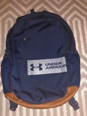 Mörkblå ryggsäck från Under Armour - Säljer en snygg mörkblå ryggsäck från Under Armour med bruna detaljer i botten och stort logotryck på framsidan. Väskan har flera fack, meshfickor på sidorna och justerbara, vadderade axelremmar. Perfekt för skola eller träning!