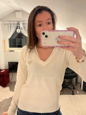 Vit v-ringad tröja med brodyr - Säljer en stilren vit v-ringad tröja med guldfärgad brodyr på bröstet. Tröjan är långärmad och har en klassisk passform med ribbade muddar. Perfekt för dig som gillar en clean och enkel look. Obs det står storlek m men passar mer som s!