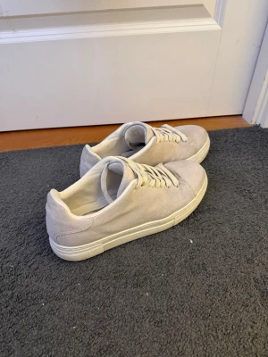 Beige sneakers i mocka med snörning - Säljer ett par stilrena beige sneakers från selected homme med platt vit sula och snörning. Skorna är tillverkade i mjuk mocka och har en rund tå. Perfekta för dig som gillar minimalistisk design och neutrala färger. Nypris 1200 kr